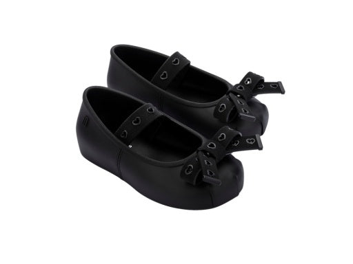 Two pairs of black matte children's Mary Jane flats with heart-cutout straps and bows on the toes.