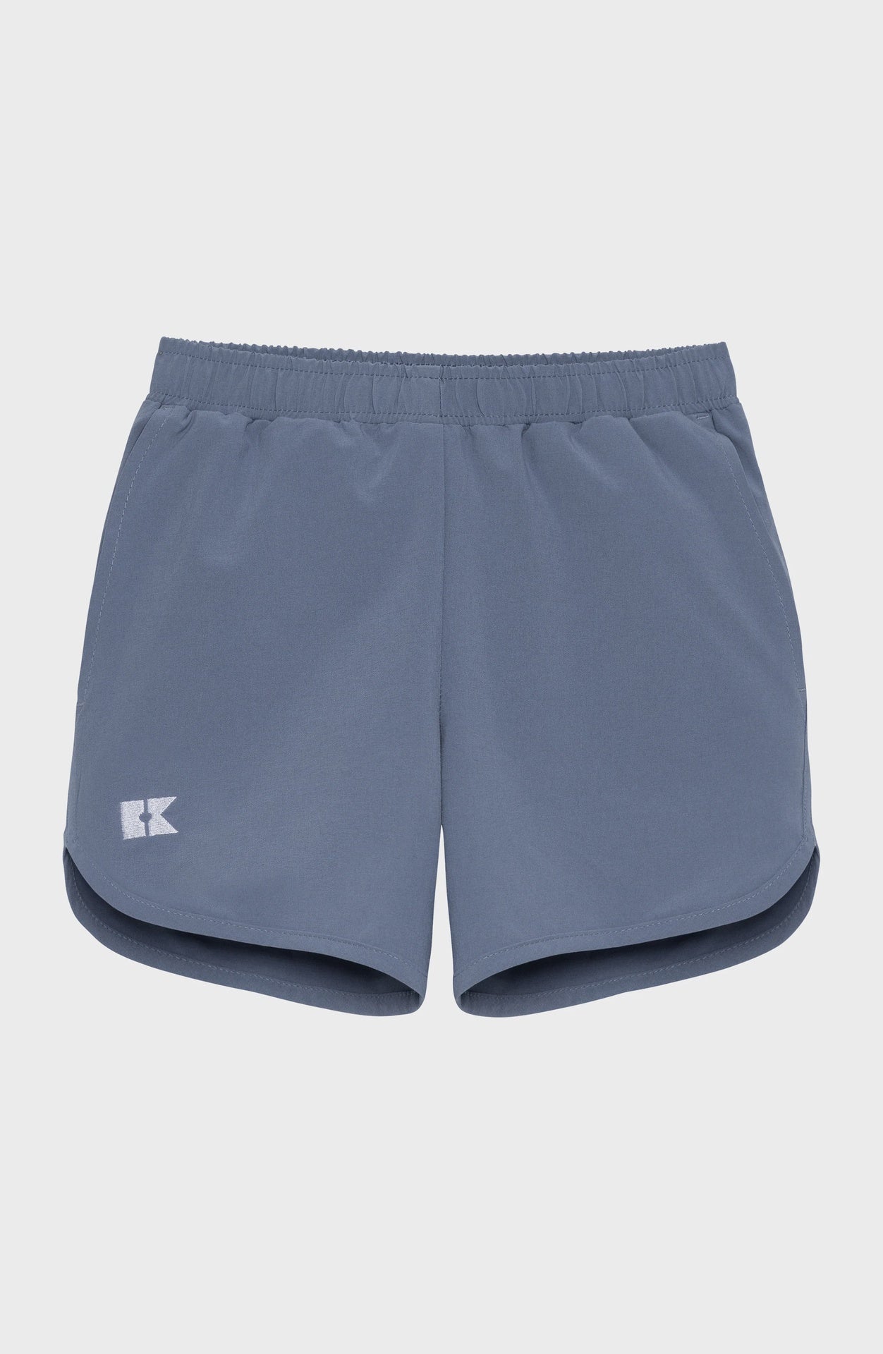 Children's slate blue athletic shorts with an elastic waistband, a white K logo on the left leg, and curved hems.