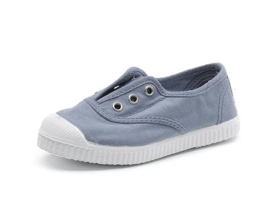 Light blue children's canvas slip-on shoe with white ribbed rubber toe cap and sole, featuring three silver eyelets.