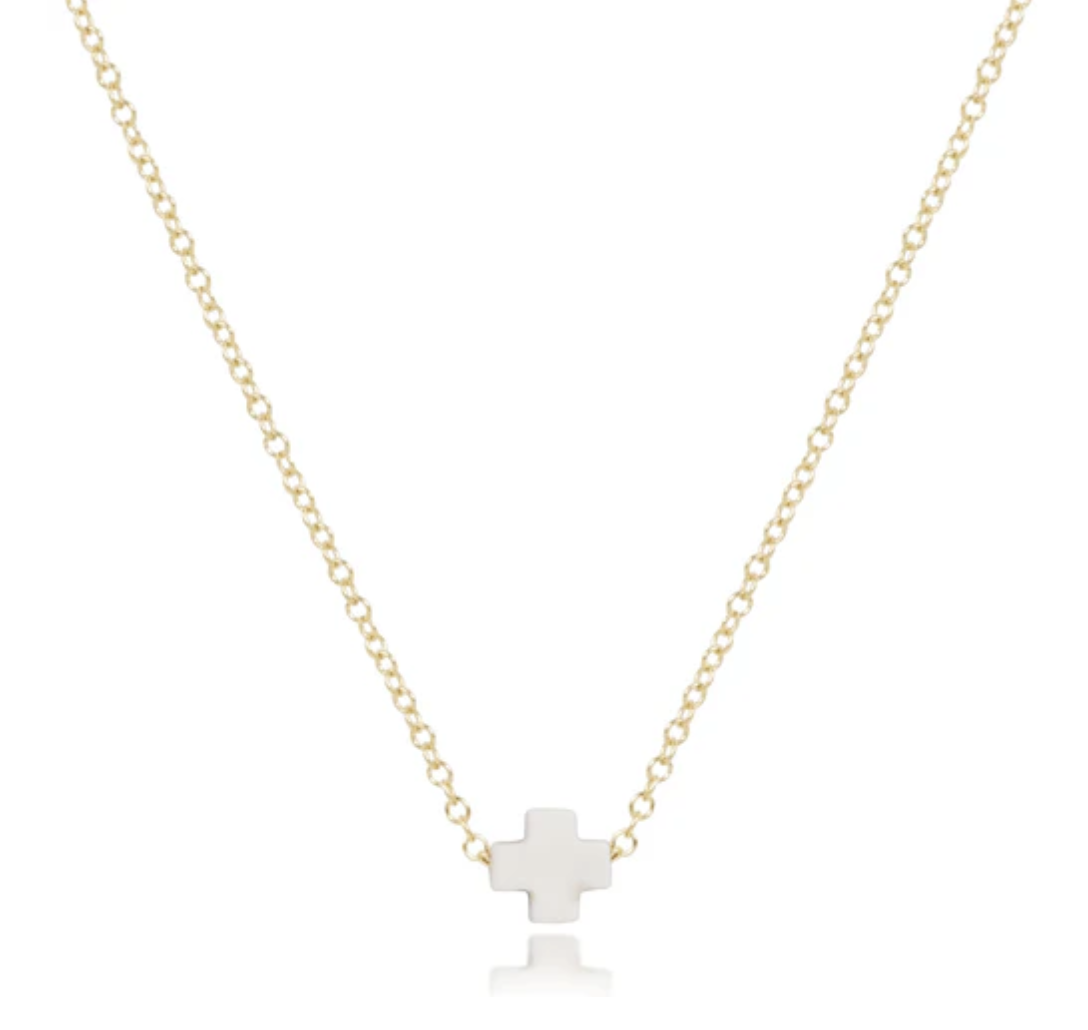 Delicate gold chain necklace with a small, flat, white cross pendant, reflected on a surface.