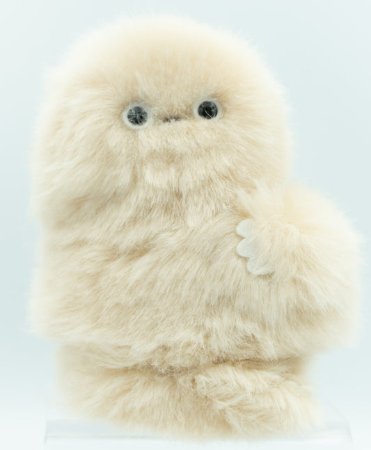 A fluffy cream-colored robotic plush toy with large black eyes and a small stitched nose, standing upright on a clear display base against a light blue background.