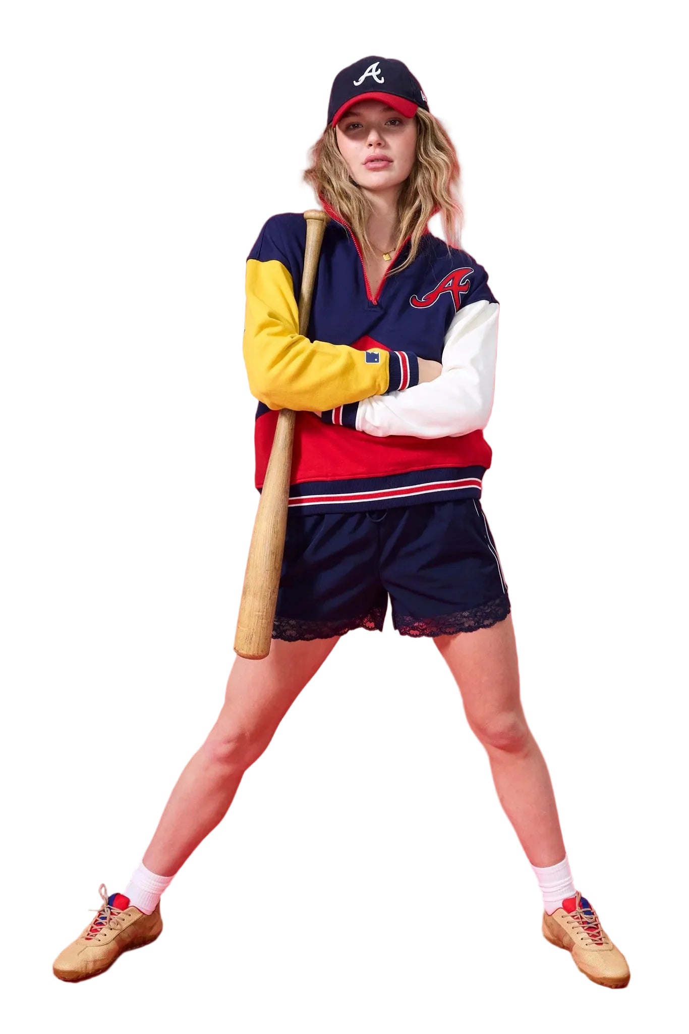 A woman modeling a vintage-style Atlanta Braves baseball uniform set, including a navy and red varsity jacket with yellow and white color-blocked sleeves, matching navy shorts, a navy cap with a red brim and white 'A' logo, white socks, and tan sneakers, while holding a wooden baseball bat against a solid red studio background.