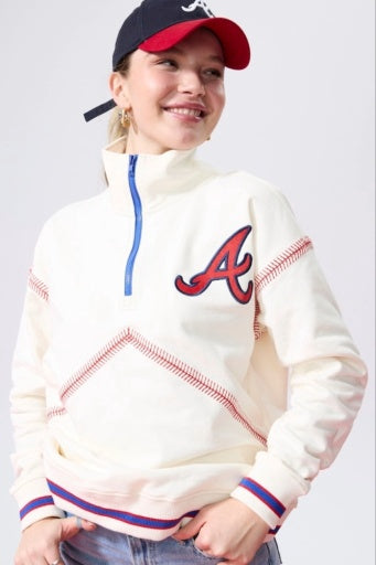 Cream quarter-zip sweatshirt featuring a red Braves 'A' logo, blue zipper, and red baseball stitching, worn by a smiling woman.